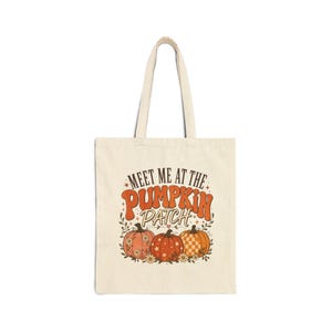 Peut inclure: Un sac fourre-tout en toile couleur crème avec une impression graphique portant le texte "MEET ME AT THE PUMPKIN PATCH" en lettres marron. Le motif comprend trois citrouilles aux tons orange et rose, ornées de motifs floraux, d'étoiles et de carreaux.
