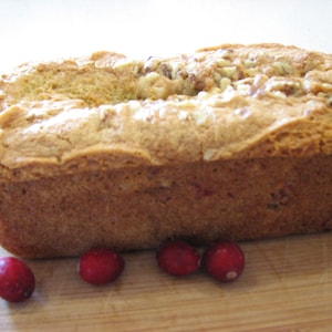 May include: A loaf of cranberry bread with white chocolate chips and walnuts on a wooden cutting board with red cranberries scattered around it.