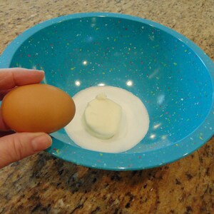May include: A blue speckled bowl with a white lump of butter and white sugar in the bottom. A brown egg is being held in a hand above the bowl.