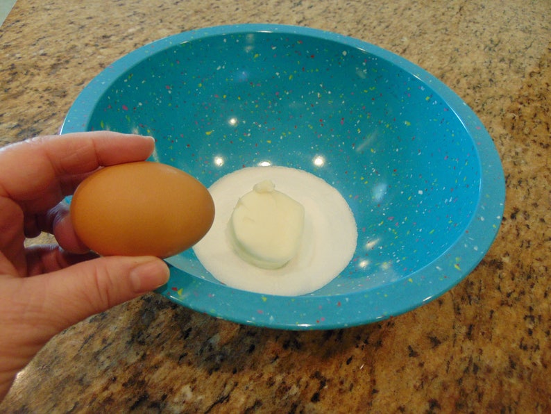 May include: A blue speckled bowl with a white lump of butter and white sugar in the bottom. A brown egg is being held in a hand above the bowl.