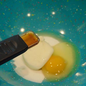 May include: A blue bowl with white specks contains a white egg yolk, a clear egg white, and a small amount of brown liquid in a clear measuring spoon.