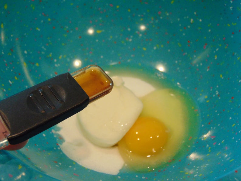 May include: A blue bowl with white specks contains a white egg yolk, a clear egg white, and a small amount of brown liquid in a clear measuring spoon.
