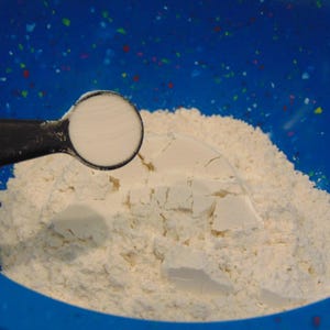May include: A blue bowl filled with white flour and a measuring spoon of baking powder.