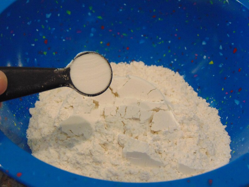 May include: A blue bowl filled with white flour and a measuring spoon of baking powder.