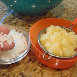 May include: A clear glass bowl filled with shredded coconut, a small amount of coconut is being held in a hand, and a strainer filled with diced pineapple sits in an orange bowl.