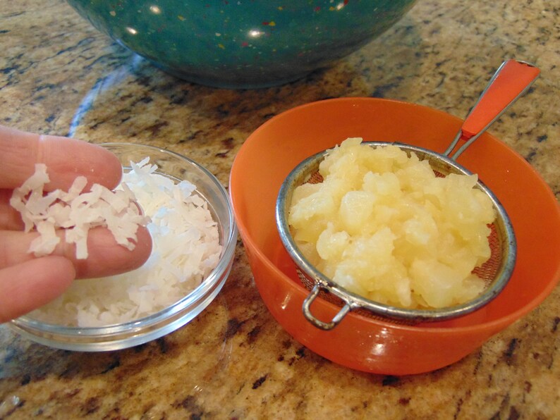 May include: A clear glass bowl filled with shredded coconut, a small amount of coconut is being held in a hand, and a strainer filled with diced pineapple sits in an orange bowl.