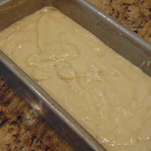 May include: A close-up of a loaf pan filled with a light brown batter. The batter is smooth and creamy, with a few swirls and bubbles on the surface.