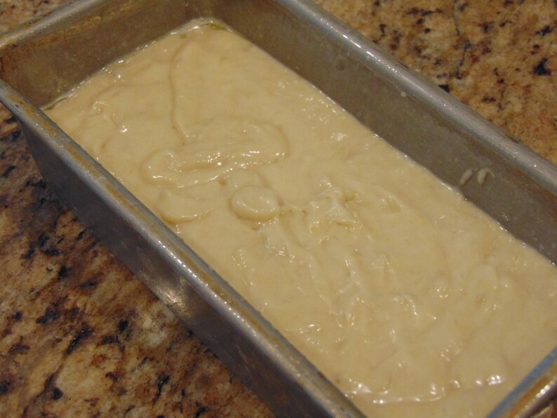 May include: A close-up of a loaf pan filled with a light brown batter. The batter is smooth and creamy, with a few swirls and bubbles on the surface.