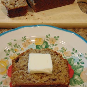 May include: A slice of banana bread with a pat of butter on top, on a white plate with a floral pattern.