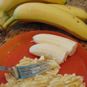 May include: A close-up of a fork and a plate of sliced bananas. The bananas are arranged in a circular pattern on the orange plate. The fork is resting on top of the bananas.