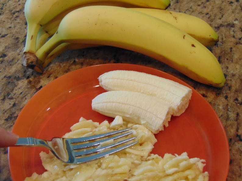 May include: A close-up of a fork and a plate of sliced bananas. The bananas are arranged in a circular pattern on the orange plate. The fork is resting on top of the bananas.