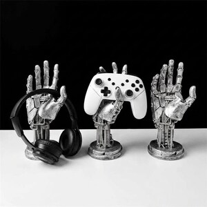May include: Three silver-colored robotic hand stands with intricate mechanical detailing. One hand holds a pair of black over-ear headphones, another holds a white video game controller, and the third hand is empty, all mounted on circular bases.