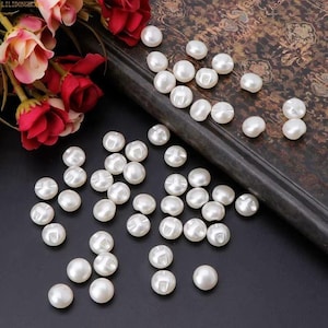 May include: A collection of small, round, white pearl-like buttons with a smooth, glossy finish. Each button features a central shank for attachment. The buttons are scattered across a dark surface next to a dark, patterned book and red flowers.