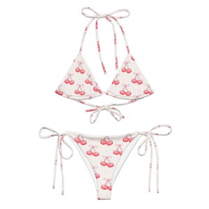 May include: A two-piece bikini set featuring a white base with a repeating pattern of small red cherries and light pink stems. The top is a triangle style with thin tie straps, and the bottom is a side-tie bikini brief with matching cherry print.