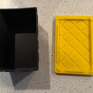 May include: A rectangular black storage container with an open top and a separate bright yellow lid. The lid features a raised diamond grid pattern on its surface, designed to fit securely onto the black base of the storage bin.