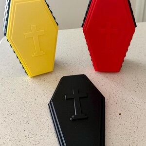 Op de afbeelding: Drie doodskistvormige bakjes in geel, rood en zwart. Elk bakje heeft een verhoogd kruis-embleem op het deksel en een zwarte basis. De gele en rode bakjes staan rechtop, terwijl het zwarte bakje plat op de ondergrond ligt.