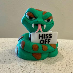 May include: A green snake figurine with brown spots, a pink forked tongue, and narrowed eyes. The snake is coiled and holds a white rectangular sign in front of its body with the black text HISS OFF printed clearly on the front surface.
