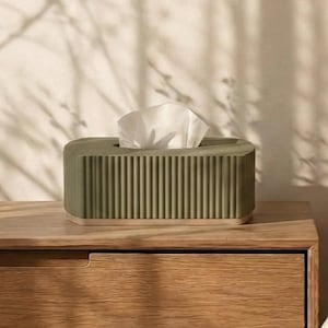 Puede incluir: Una caja para pañuelos rectangular de color verde oliva con una textura acanalada vertical en los lados y una base de madera clara lisa. Un pañuelo blanco asoma por la abertura ovalada situada en la parte superior del recipiente.