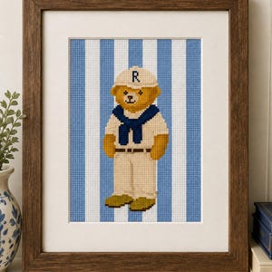 May include: A framed cross-stitch embroidery featuring a golden-brown teddy bear wearing a cream-colored baseball cap with the letter R, a dark blue sweater draped over the shoulders, and cream-colored pants, set against a blue and white striped background.