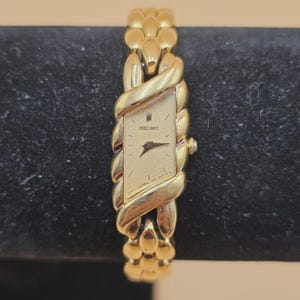May include: A gold-tone Seiko wristwatch featuring a diamond-shaped face with a textured gold dial, thin black hands, and a unique braided-style metal link bracelet. The watch face displays the Seiko logo and text indicating it is made in Japan.