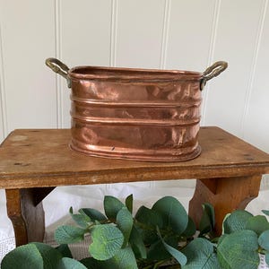 May include: An oval copper planter with horizontal ribbed detailing and two ornate brass handles on the sides. The metallic surface shows a polished finish with natural variations in colour and texture across the exterior body.