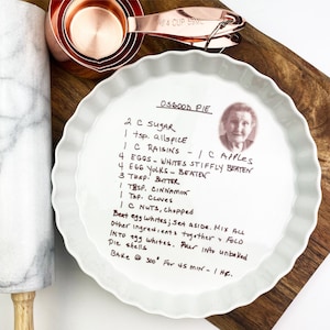 May include: A white pie plate with a handwritten recipe for Osgood Pie. The recipe includes ingredients like sugar, allspice, raisins, apples, eggs, butter, cinnamon, cloves, and nuts. The recipe also includes instructions for baking the pie at 300 degrees Fahrenheit for 45 minutes.
