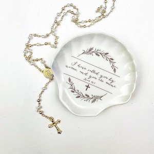 May include: White ceramic dish with a gold rim and a floral design. The dish has the text "I have called you by name, and you are mine.  John 10:3" printed on it. A gold and white rosary necklace is lying on a white surface next to the dish.