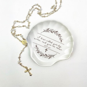May include: White ceramic dish with a gold rim and a floral design. The dish has the text "I have called you by name, and you are mine.  John 10:3" printed on it. A gold and white rosary necklace is lying on a white surface next to the dish.