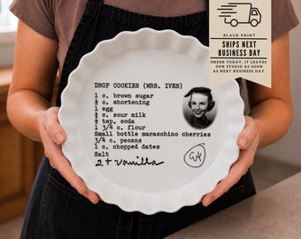 Könnte beinhalten: Eine weiße, gewellte Keramik-Kuchenform mit einem aufgedruckten Rezept für Drop Cookies (Mrs. Ives) in schwarzer Schreibmaschinenschrift. Das Design enthält ein kleines Schwarz-Weiß-Porträt einer Frau, eine handschriftliche Notiz für Vanille und eine eingekreiste Signatur.