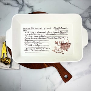 May include: White ceramic baking dish with a handwritten recipe for Broccoli Salad (Delicious) on the front. The recipe includes ingredients like broccoli, bacon, red onion, mayonnaise, sugar, and vinegar. The dish has a brown floral design with a basket of apples and a pie.