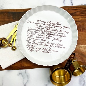 May include: A white pie plate with a handwritten recipe for Mom's Cherry Pie. The recipe includes ingredients like self-rising flour, shortening, and cold water. It also mentions crimping the edges and baking at 400 degrees Fahrenheit for 20 minutes.