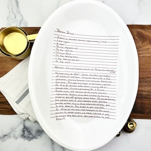 May include: A white ceramic oval platter with a handwritten recipe for banana bread. The recipe includes ingredients like bananas, sugar, eggs, vanilla, flour, baking soda, baking powder, and salt. Optional ingredients include chopped walnuts or chocolate chips. The recipe also includes instructions for preheating the oven, preparing the loaf pan, and baking the bread.