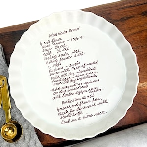 May include: A white ceramic pie plate with a handwritten recipe for Irish Soda Bread. The recipe includes ingredients like flour, sugar, baking soda, buttermilk, and raisins. The plate has a scalloped edge and is sitting on a wooden surface.