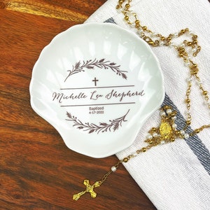 May include: A white, shell-shaped ceramic dish with a personalized inscription and cross design. The text reads "Michelle Lea Shepherd Baptized 4-17-2022". A gold rosary with pearl beads rests on a white and blue striped cloth.