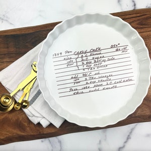 May include: A white ceramic pie plate with a handwritten recipe for "Crazy Cake" on it. The recipe includes ingredients like flour, sugar, baking soda, salt, cocoa, oil, vinegar, vanilla, and cold water. The plate is 13x9 inches and the baking temperature is 350 degrees Fahrenheit for 30-45 minutes.