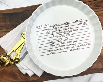 May include: A white ceramic pie plate with a handwritten recipe for "Crazy Cake" on it. The recipe includes ingredients like flour, sugar, baking soda, salt, cocoa, oil, vinegar, vanilla, and cold water. The plate is 13x9 inches and the baking temperature is 350 degrees Fahrenheit for 30-45 minutes.