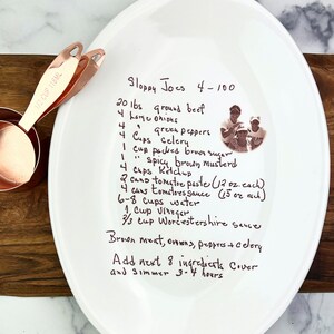 May include: A white ceramic plate with a handwritten recipe for Sloppy Joes for 4-100 people. The recipe includes ingredients like ground beef, onions, green peppers, celery, brown sugar, brown mustard, ketchup, tomato paste, tomato sauce, water, vinegar, and Worcestershire sauce. The recipe also includes instructions to brown the meat, onions, peppers, and celery, then add the remaining ingredients and simmer for 3-4 hours.