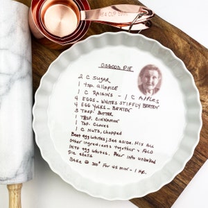 May include: A white ceramic fluted pie dish featuring a handwritten Osgood Pie recipe and a small portrait of an elderly person. The dish is accompanied by copper measuring cups and a marble rolling pin, all arranged on a wooden surface.