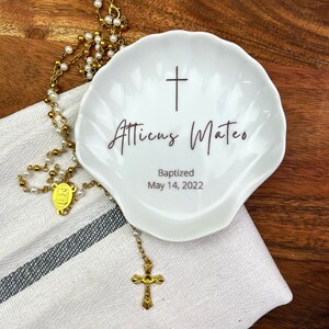 May include: White ceramic seashell dish with a cross and the text "Atticus Mateo Baptized May 14, 2022". A gold rosary with a cross and a medal is on a white cloth next to the dish.
