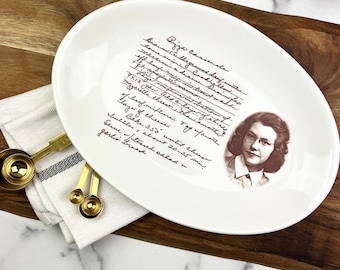 May include: An oval white ceramic serving platter with a handwritten recipe and a sepia-toned portrait of a woman. The recipe is in black ink. Gold measuring spoons and a white cloth are on the side.