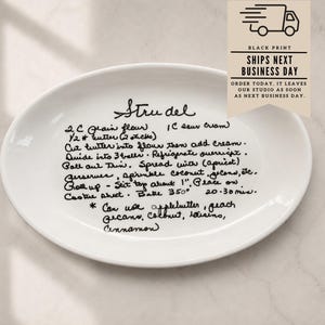 May include: A white oval ceramic platter featuring a black handwritten recipe for Strudel. The text includes ingredients like flour, butter, and sour cream, along with baking instructions. The surface is smooth and glossy with clear, legible black script.