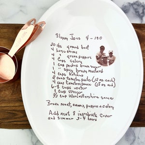 May include: A white ceramic plate with a handwritten recipe for Sloppy Joes for 4-100 people. The recipe includes ingredients like ground beef, onions, green peppers, celery, brown sugar, brown mustard, ketchup, tomato paste, tomato sauce, water, vinegar, and Worcestershire sauce. The recipe also includes instructions to brown the meat, onions, peppers, and celery, then add the remaining ingredients and simmer for 3-4 hours.