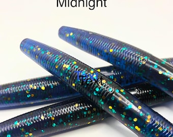 May include: A collection of soft plastic fishing lures in a dark blue color with embedded gold and teal glitter. The cylindrical lures feature a ribbed texture along the body. The word Midnight is printed in black text above the lures.