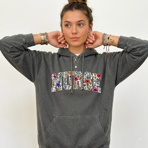 Könnte beinhalten: Ein anthrazitfarbener Kapuzenpullover mit dem Wort NURSE, das in einer fetten Blockschrift gedruckt ist. Die Buchstaben sind mit einem farbenfrohen, detaillierten Blumenmuster aus verschiedenen kleinen Blüten und grünen Blättern gefüllt.