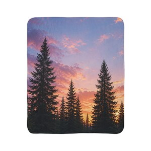 May include: A rectangular throw blanket featuring a printed landscape of dark, silhouetted evergreen trees against a vibrant sunset sky filled with soft pink, orange, and purple clouds. The blanket has rounded corners and a smooth, uniform texture.