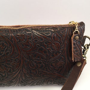 May include: A brown leather clutch with an embossed floral pattern and a gold zipper. The clutch has a detachable strap with a gold chain and a brown leather tassel.