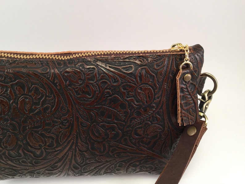May include: A brown leather clutch with an embossed floral pattern and a gold zipper. The clutch has a detachable strap with a gold chain and a brown leather tassel.