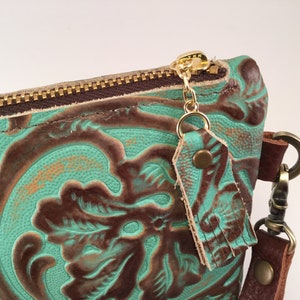 May include: A teal and brown leather clutch with a gold zipper and a tassel. The clutch has a floral embossed design.