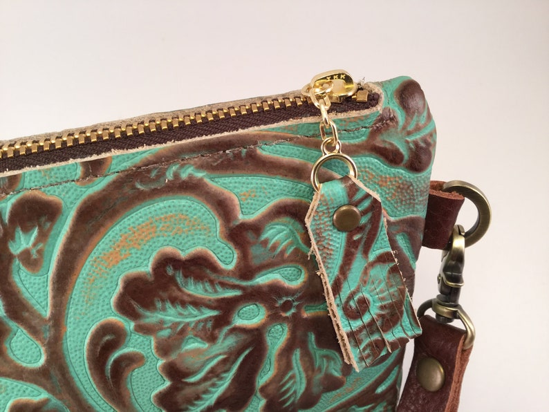 May include: A teal and brown leather clutch with a gold zipper and a tassel. The clutch has a floral embossed design.