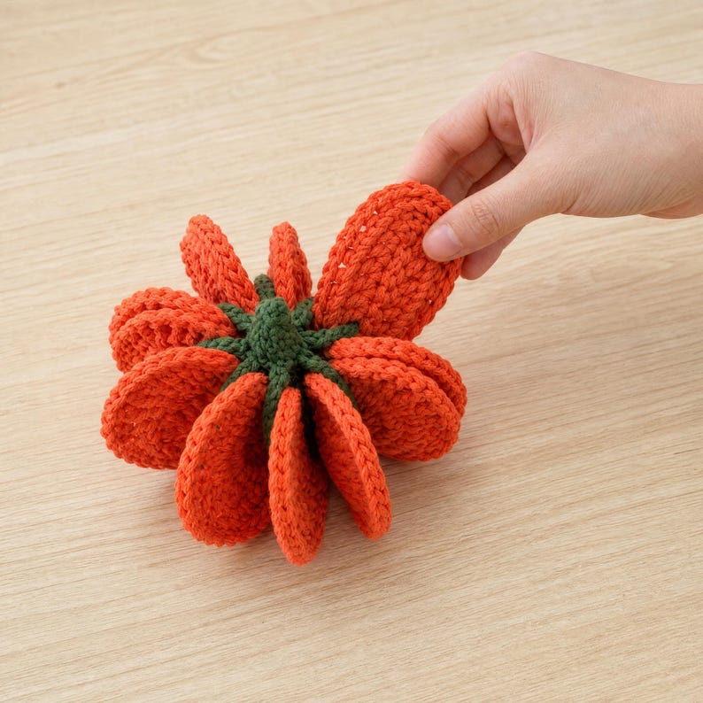 May include: A crocheted pumpkin decoration featuring multiple vibrant orange, rounded petal-like segments arranged in a circular pattern around a central dark green stem. The textured yarn construction creates a layered, dimensional appearance.